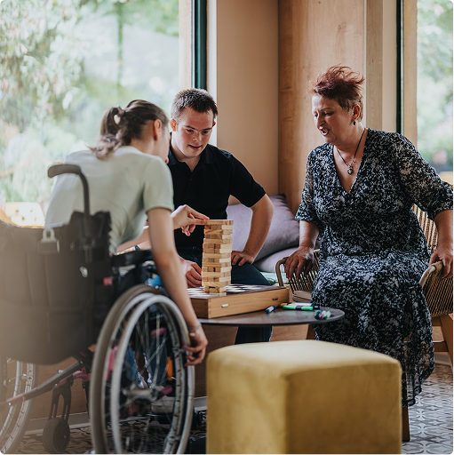 Trois personnes, dont une en fauteuil roulant et une autre atteinte de trisomie 21, jouant ensemble à un jeu de Jenga dans un espace convivial.
