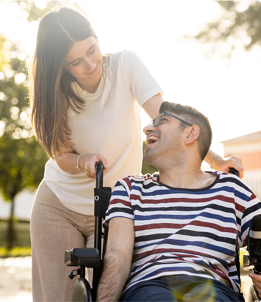 Jeune femme poussant un homme en fauteuil roulant, échangeant un sourire complice dans un parc ensoleillé.